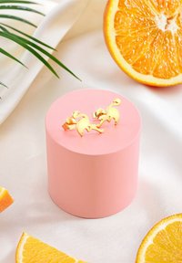 Boucles d'oreilles flamant rose en or sur un cylindre de présentation rose. Autour, des tranches d'orange et une feuille verte, le tout sur un fond en tissu clair.