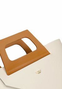 Strukturalna torba tote w kremowym kolorze z brązowym uchwytem i prostokątnym wycięciem. Złoty detal z logo z przodu. Gładka tekstura.