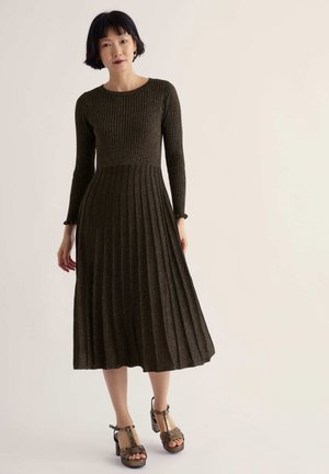 Femme portant une robe midi en maille côtelée vert foncé avec manches longues et jupe plissée, associée à des sandales marron à talons hauts et à brides.