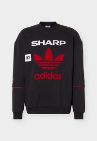 Μαύρο φούτερ με λευκό κείμενο "SHARP", κόκκινο λογότυπο Adidas trefoil, κείμενο "adidas" και ένα μικρό λευκό έμβλημα με το "92" στην αριστερή περιοχή του στήθους.