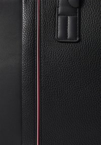 Surface en cuir noir texturé avec une bande en cuir lisse et un détail étroit de bandes verticales rouges, blanches et bleues à côté d'une languette en cuir cousue.