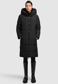 Zwarte pufferjas met een capuchon, voorzien van een ritsluiting, gewatteerd ontwerp en zijzakken. Gekozen met zwarte legging en kniehoge laarzen.