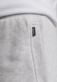 Primer plano de pantalones de chándal gris claro con una cinturilla acanalada, un bolsillo lateral y una pequeña etiqueta negra que dice "SUPERDRY" en la costura del bolsillo.