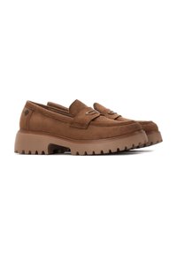 Mocasines de ante marrón con una suela gruesa y estriada. Presenta una correa decorativa en el empeine y un sutil detalle del logo en el lateral.