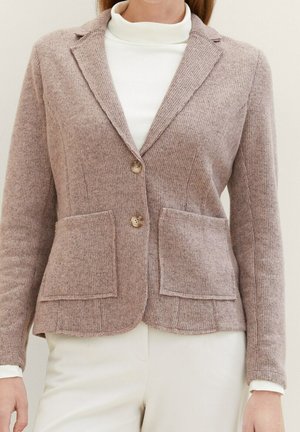 Woman wearing a beige knit blazer with two front pockets and buttons, layered over a white turtleneck and paired with white pants.