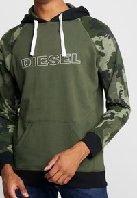 Oliwkowa zielona bluza z kapturem w czarnym kolorze, białymi sznurkami i rękawami w kamuflażu. Posiada przednią kieszeń oraz duże logo „DIESEL” w białym kolorze.