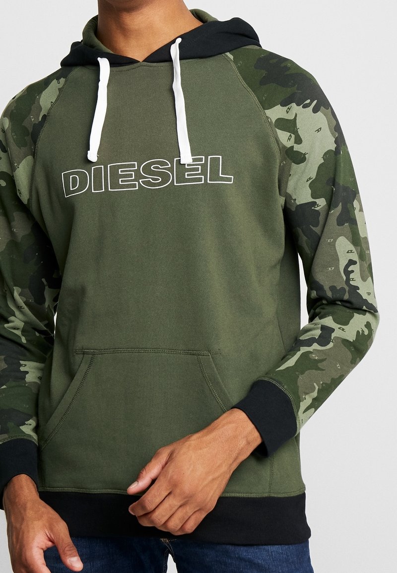 Oliwkowa zielona bluza z kapturem w czarnym kolorze, białymi sznurkami i rękawami w kamuflażu. Posiada przednią kieszeń oraz duże logo „DIESEL” w białym kolorze.