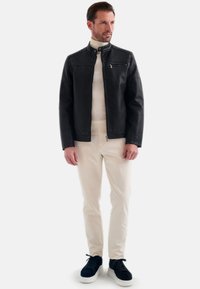 Zwarte leren jasje met een staande kraag, ritszak en een zachte textuur, gecombineerd met een crèmekleurige trui en lichte broek, gedragen door een mannelijke model.