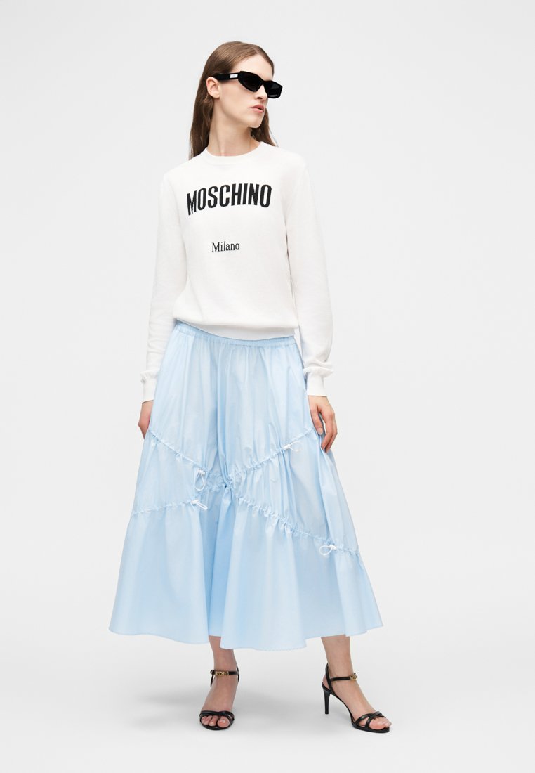 Femme portant un pull Moschino blanc, une jupe midi bleu clair à volants, des lunettes de soleil noires et des sandales à talons hauts noires, debout devant un fond blanc.
