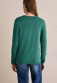 Grüner, langärmeliger Strickpullover mit Rundhalsausschnitt und geripptem Saum. Weiche Textur und einfarbig, kombiniert mit blauen Jeans.