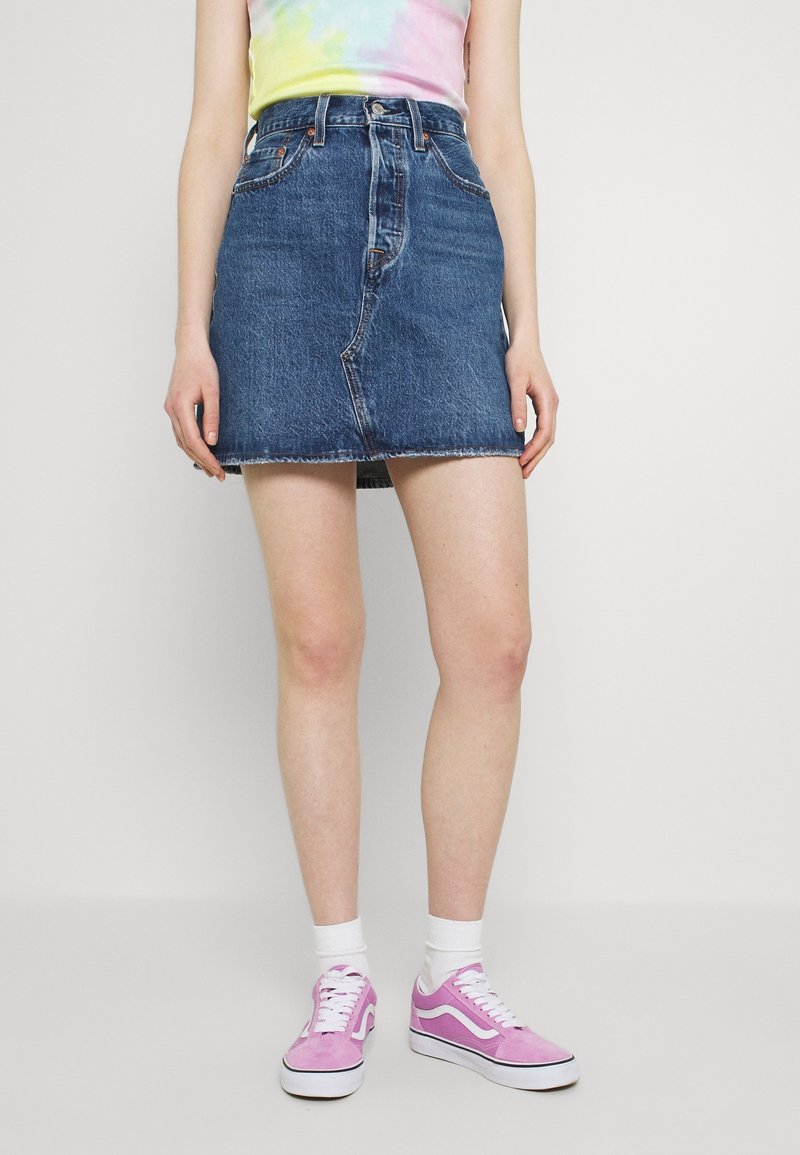 Personne portant une mini-jupe en jean bleu, des chaussettes blanches, des baskets roses et un haut tie-dye pastel sur un fond uni.