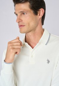 Polo shirt bianco a maniche lunghe con logo ricamato argento sul petto. Presenta un colletto classico e accenti a righe blu su polsini.