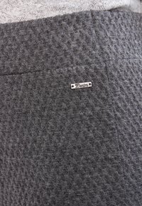 Tessuto grigio testurizzato con un motivo a diamante, caratterizzato da un piccolo etichetta metallica che reca la scritta "Denim". L'area è liscia con una leggera lucentezza.