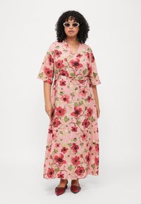 Floral maxi dress featuring a pink background with red and green poppy patterns, short sleeves, and a cinched waist. Worn with glasses and heels.
