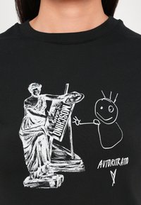 Svart t-skjorte med et hvitt grafisk bilde av en statue som holder et skilt, ved siden av en strekfigur-tegning. Teksten sier "AUTORITRATTO."