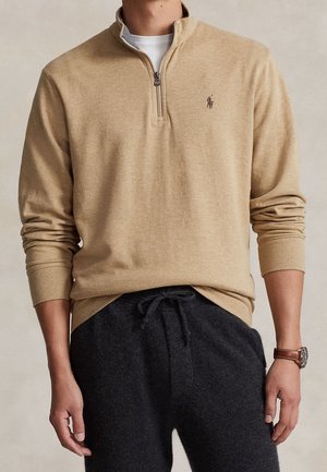Beiger Viertel-Reißverschluss-Pullover mit einem kleinen gestickten Logo auf der Brust, kombiniert mit dunklen Kordelzughosen und einer braunen Lederarmbanduhr.