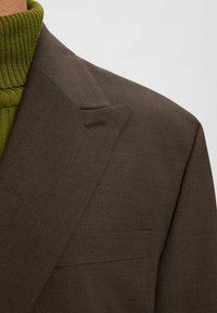 Pruun jakk, millel on sile tekstuur, esineb sälguga reväärid ja rinnatasku. Selle all paistab soonikuline roheline poolkaelusega särk.