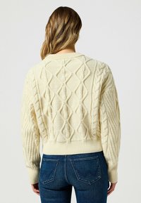 Maglione a trama cable beige con un motivo a rombo testurizzato e orlo a costine, abbinato a jeans blu scuro. Il design sul retro mette in evidenza cuciture uniche.