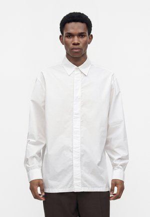Jeune homme portant une chemise blanche ample boutonnée à manches longues et un pantalon foncé, debout devant un fond blanc uni.