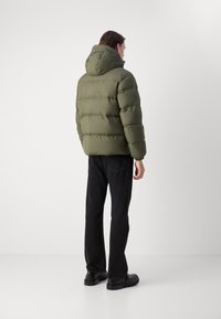 Olivegrüner Puffer-Jacke mit horizontaler Steppung, Kapuze und elastischen Bündchen, kombiniert mit schwarzen Jeans und schwarzen Schuhen.