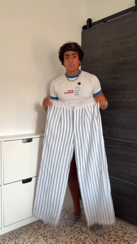 Junger Mann in einem weißen T-Shirt, der eine Hose mit blauen vertikalen Streifen in der Hand hält, steht in einem Raum mit einer Holztür und einem gefliesten Boden.
