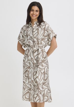 Naeratav naine, kes kannab beeži ja valge mustriga vööga varustatud lühikeste varrukatega särkkleiti, seisab ühevärvilise tausta ees.