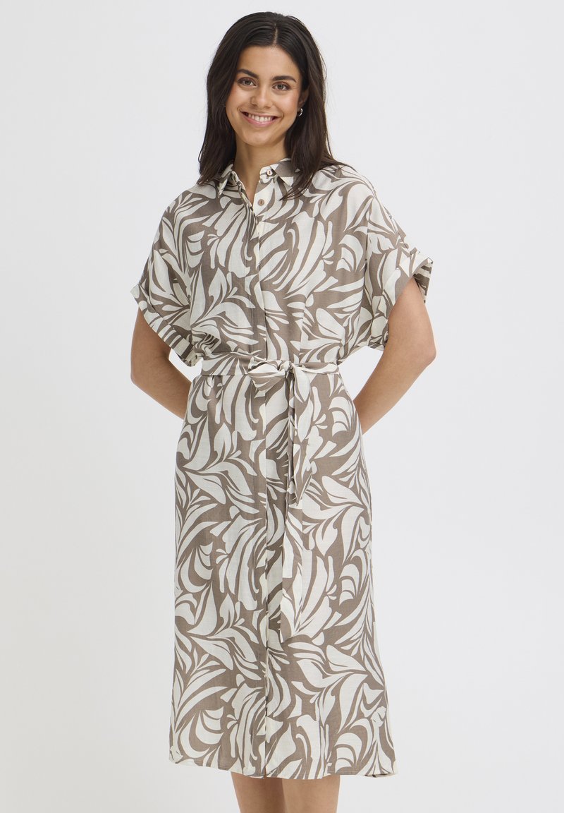 Usmievavá žena v béžovo-bielej vzorovanej košilovej šaty s opaskom a krátkymi rukávmi stojaca pred jednoduchým pozadím.