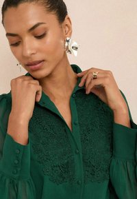 Blusa verde con ricami floreali sul petto, chiusura a bottoni e maniche a sbuffo. Orecchini astratti in argento e un anello d'oro completano il look.