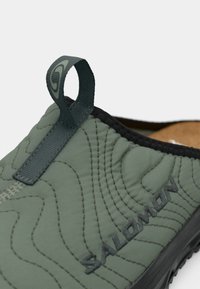 Grønn polstret slip-on sko med kviltet mønster, svarte detaljer og en trekkflik. Har en teksturert såle og "Salomon"-merke.