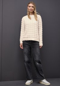 Suéter de punto crema con patrón texturizado y ondulado; combinado con jeans oscuros sueltos y zapatillas blancas; fondo gris.