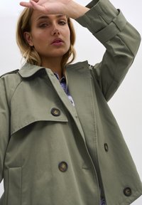 Trench coat color verde oliva in tessuto leggero con grandi bottoni neri, tasche laterali e un colletto pronunciato. Design su misura con vestibilità ampia.