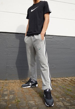 Persona che indossa una t-shirt nera Nike, pantaloni della tuta grigi Nike con strisce laterali e sneakers nere Nike, in piedi davanti a un muro bicolore.