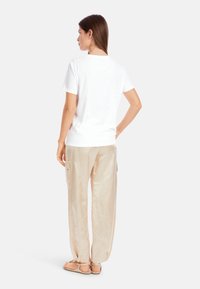 Weißes Kurzarmhemd kombiniert mit hellbeigen, glänzenden, weiten Hosen mit Taschen, wobei das Modell von hinten gesehen wird.
