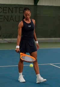 Tennis player in a fitted black tank top and skirt, holding an orange tennis racket. Green court background with a tennis ball in motion.