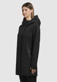 Veste noire imperméable avec capuche, dotée d'une fermeture éclair sur le devant, de deux poches latérales et d'une coupe droite. Texture lisse et mate.