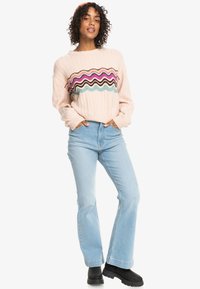 Maglione a coste rosa chiaro con design zigzag multicolore sul petto, abbinato a jeans svasati blu chiaro e stivali chunky neri.