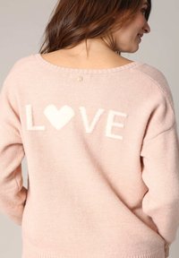 Pull léger rose avec le texte "LOVE" blanc et un cœur. Présente un col large et des poignets côtelés. Tissu doux et texturé avec un petit accent doré.