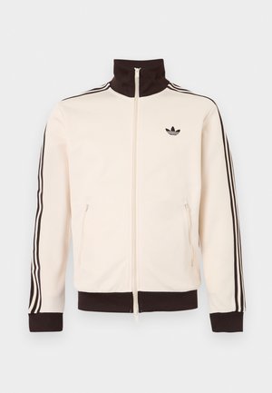 Veste de survêtement crème avec col, poignets et ourlet noirs, présentant un design à trois bandes noires sur les manches et un logo Adidas noir sur la poitrine.
