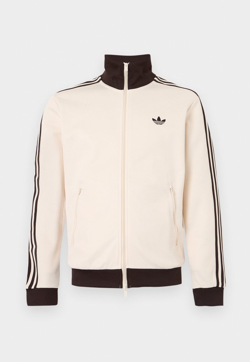 Chaqueta de chándal color crema con cuello, puños y dobladillo negros, presenta un diseño de tres rayas negras en las mangas y un logo negro de Adidas en el pecho.
