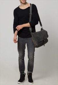 Chemise noire à manches longues avec un col usé, jeans gris, baskets noires et un sac messager en cuir gris avec rabat et boucle.