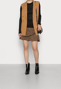 Hiekansävyinen hihaton bleiseri mustan ribbineuletopin päällä, yhdistetty kuvioituun ruskeaan minihameeseen. Viimeistelty mustilla kiiltävillä nilkkureilla.