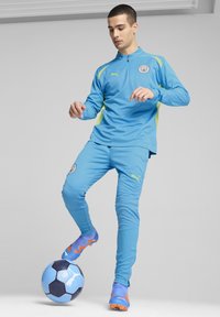 Debeszilā sporta treniņtērps ar garām piedurknēm un biksēm, ar dzelteniem akcentiem. Spēlētājs stāv uz melna un zila futbola bumbas.