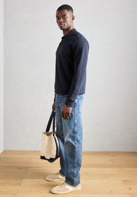 Sötétkék, hosszú ujjú polo ing, világoskék, egyenes szárú farmer, bézs sportcipő és egy bézs vászontáska sötét pántokkal. Fa padló háttér.