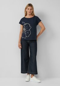 Marineblauw T-shirt met een zilveren bloemenontwerp, korte mouwen, gecombineerd met brede donkere denim jeans en witte sneakers. Gladde textuur.