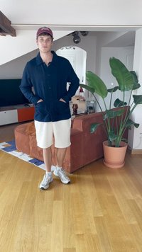 Marineblå skjorte med knapper og lommer, hvite shorts og grå sneakers. Innendørs setting med en potteplante og en brun kordfløyelssofa.
