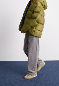 Giacca a vento verde con cappuccio, pantaloni in corduroy grigi e scarpe grigie con accenti gialli e verdi. La giacca presenta un design trapuntato.