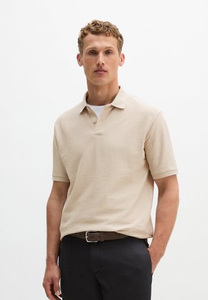 Man draagt een beige poloshirt met korte mouwen over een wit ondershirt, zwarte broek en een bruine riem, staand tegen een effen witte achtergrond.
