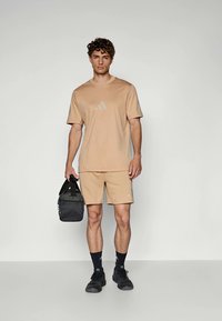 Beige sports t-skjorte og shorts, laget av lett materiale, med subtil branding. Holder en svart treningsbag, iført svarte sokker og sko.