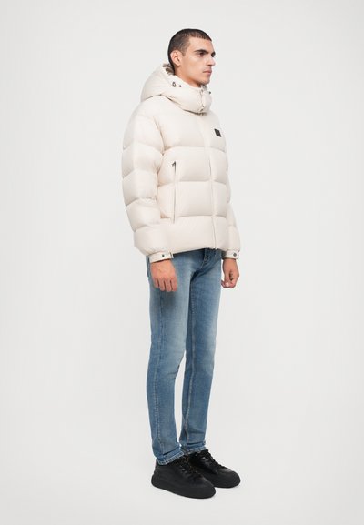 Beige foret jakke med horisontalt quiltet mønster, hætte og frontlynlås. Bæres med blå tætsiddende jeans og sorte sneakers.