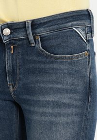 Σκούρο μπλε τζιν παντελόνι με ομαλή υφή, σχεδιασμό με πέντε τσέπες, χρυσή ραφή και μικρό λευκό ετικέτα λογότυπου κοντά στην μπροστινή τσέπη.